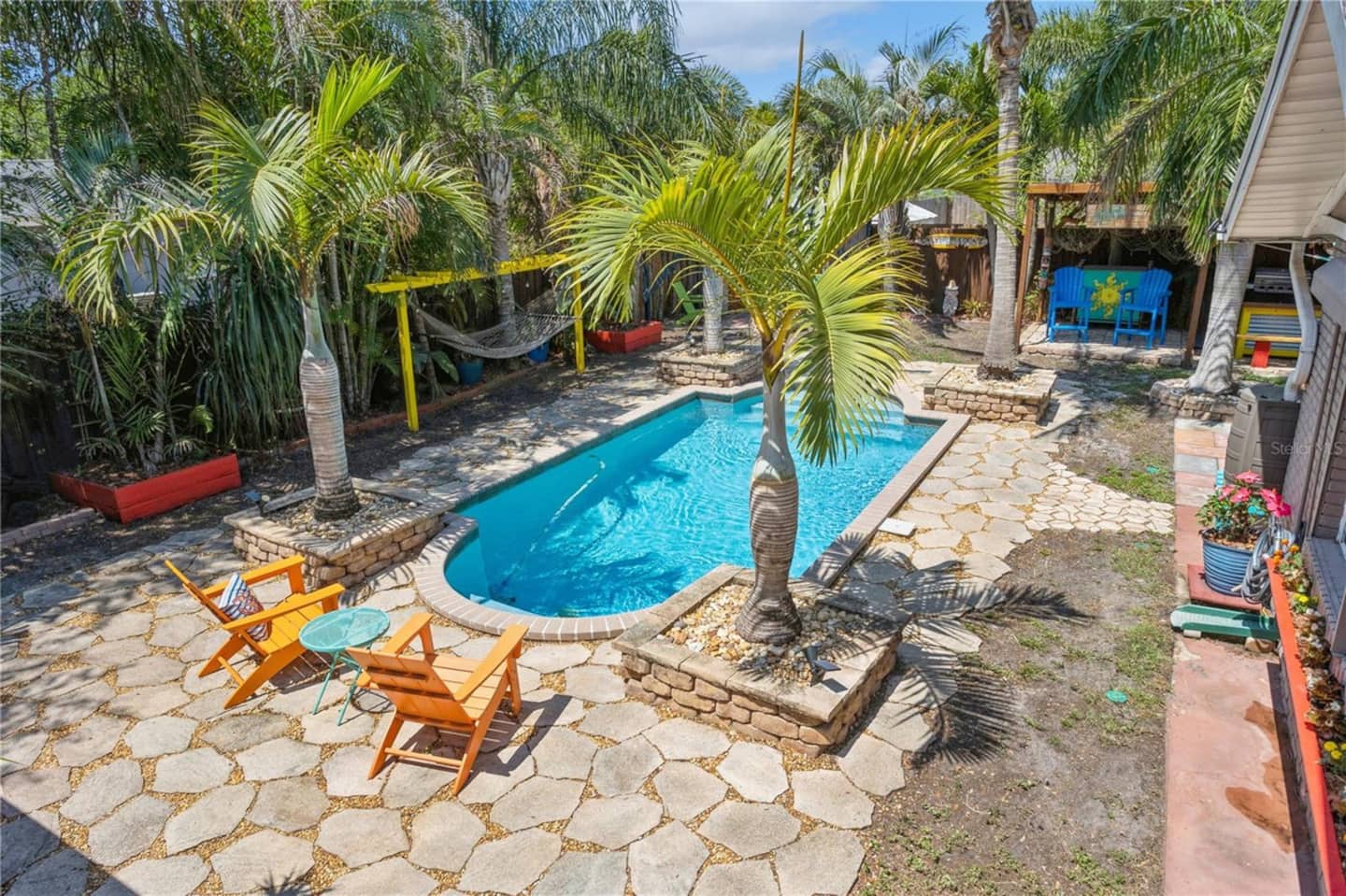 The kidney-shaped pool in daylight, ringed by palms, with orange Adirondack chairs on a flagstone deck.