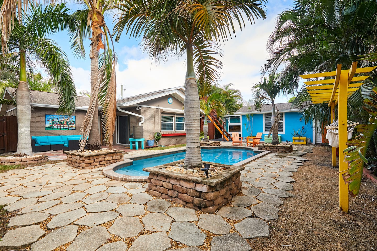 The full backyard: the single-storey house, a blue outbuilding, the pool and a yellow pergola among palms.