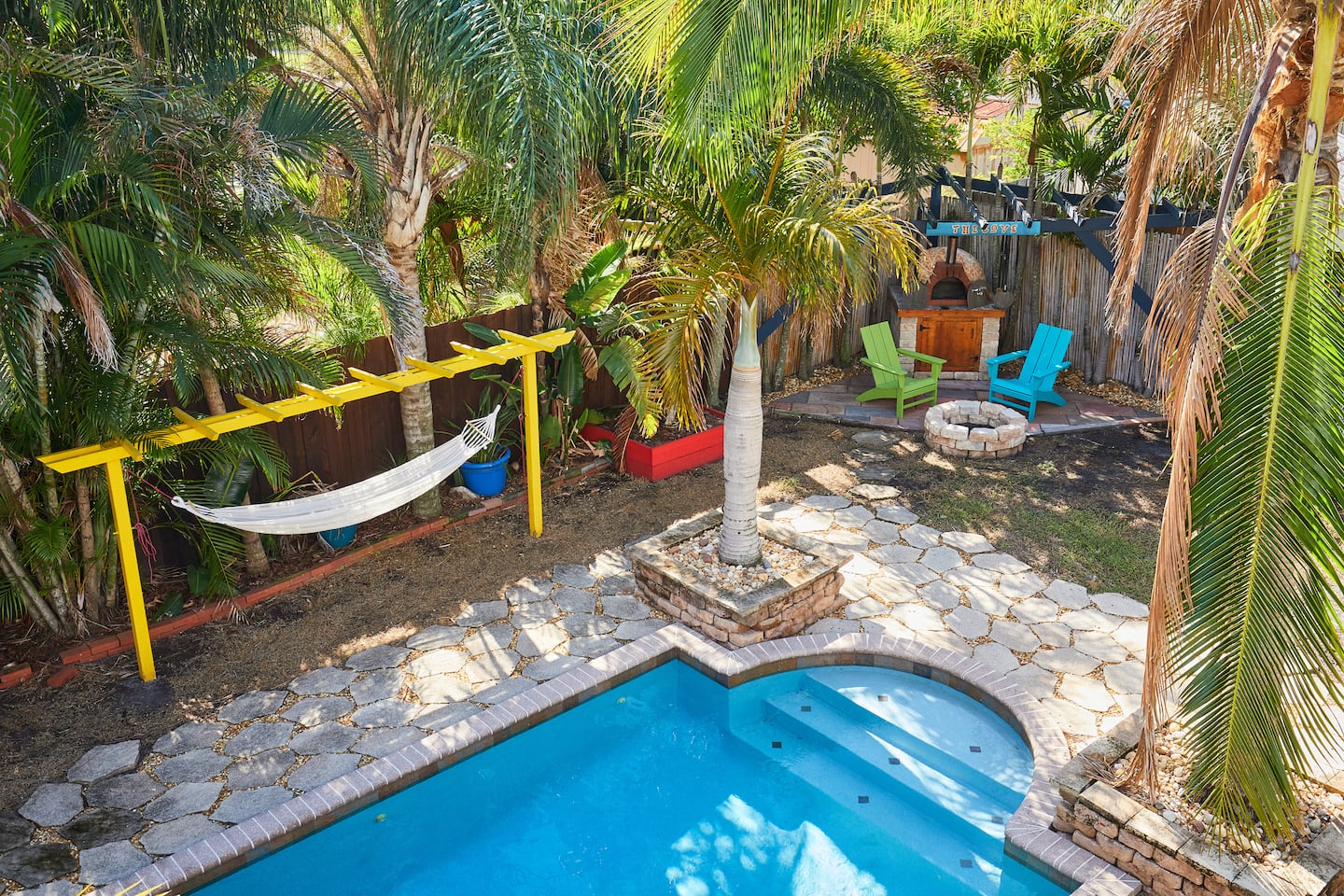 A hammock slung under a yellow pergola beside the pool, with the fire pit and chairs beyond.