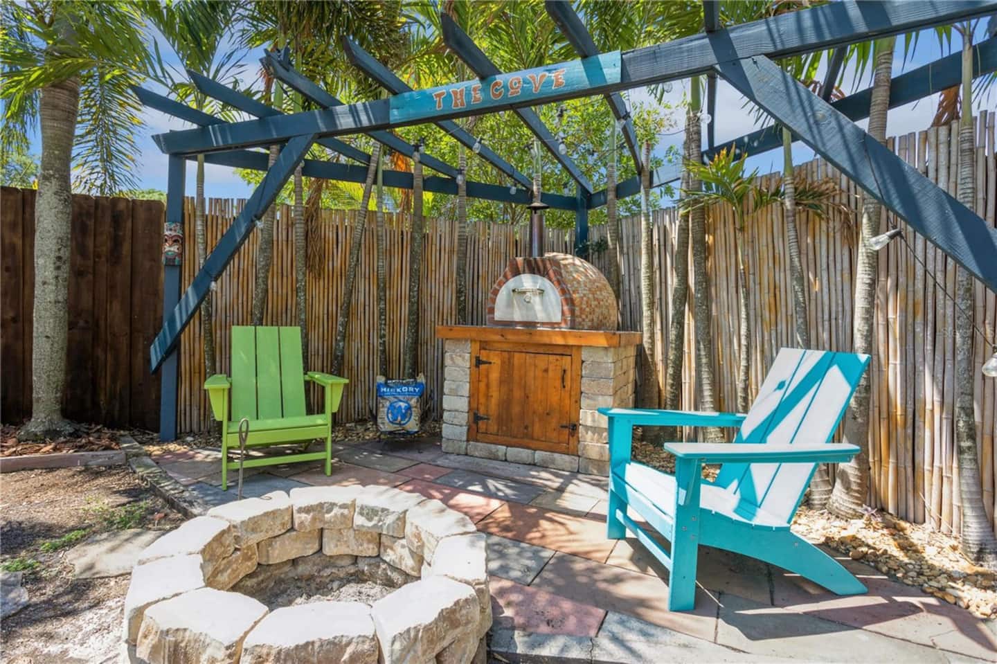 A stone fire pit and a wood-fired oven beneath a pergola signed "The Cove", against a bamboo fence.